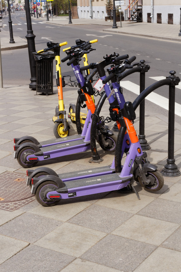 Eine Reihe von Elektro-Scootern, die auf der Straße geparkt sind, mit einem Mülleimer im Vordergrund, Gebäuden, Bäumen, Pfosten, Schildern und Fahrzeugen im Hintergrund.