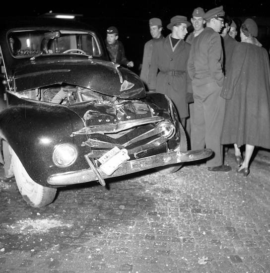 Schwarz-weiß-Bild eines schwer beschädigten Autos am Straßenrand mit eingedellter Motorhaube und zersplitterten Fenstern, umgeben von einer Gruppe von Menschen, die die Szene begutachten.