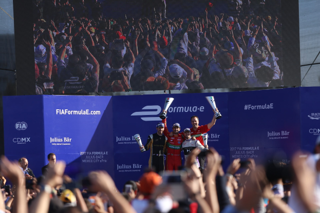 Gruppe von Menschen auf einem Formel-E-Podium in Mexiko-Stadt mit Pokale, Sponsorenplakaten und einem Bildschirm im Hintergrund.