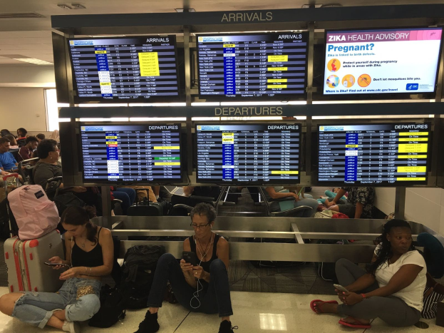 Menschen sitzen auf dem Boden eines Flughafens, einige halten Handys, mit Taschen drumherum und Zika-Virus-Informationen auf Bildschirmen im Hintergrund.