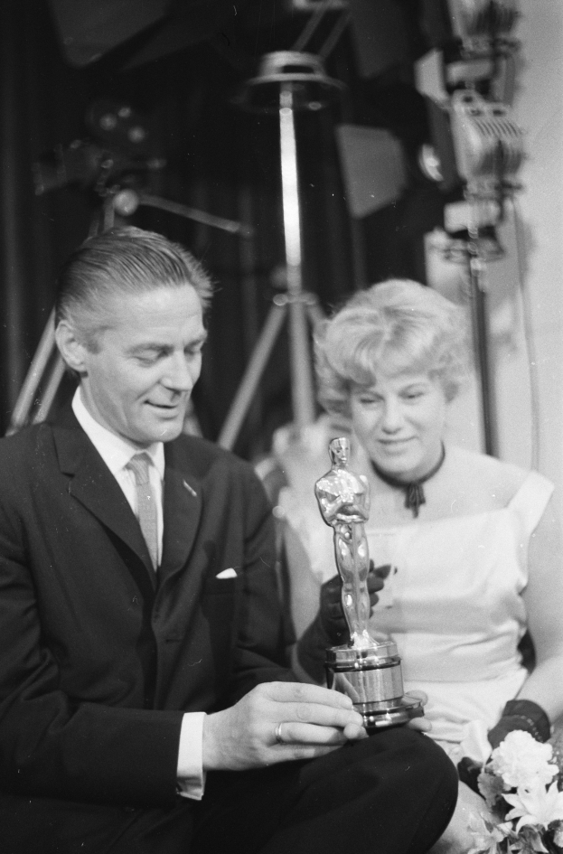 Ein Mann in einem schwarzen Blazer, einem weißen Hemd und einer Krawatte hölt eine Oscar-Statue in der Hand, waehrend er neben einer Frau in Abendgarderobe sitzt, die einen Blumenstrau├č hölt, mit weicher Beleuchtung im Hintergrund.