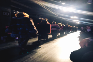 Eine Gruppe von Menschen läuft auf einem Laufsteg bei einer Fashion Show, beleuchtet von hellen Scheinwerfern, mit Zuschauern auf beiden Seiten.