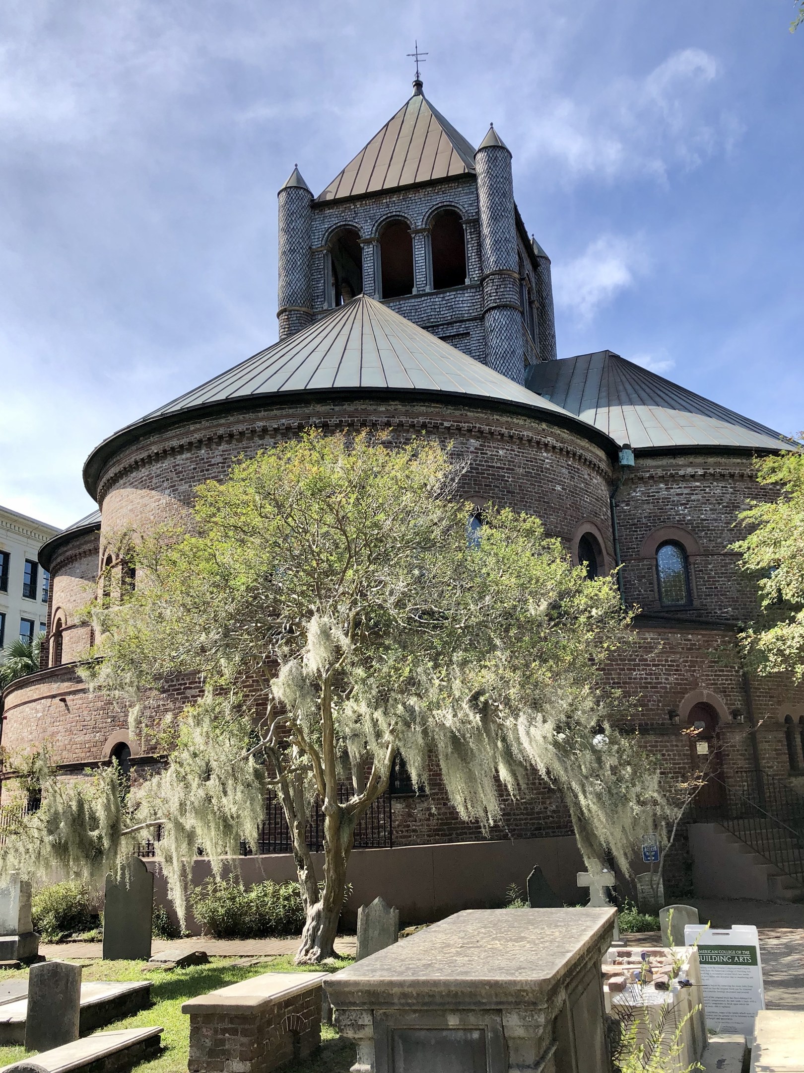 Außenansicht der Kirche des Heiligen Grabes in Charleston, South Carolina, mit einem Turm, einem Baum, umgeben von Gebäuden, einer Treppe und einem bewölkten Himmel, mit Gras und einer Texttafel unten.