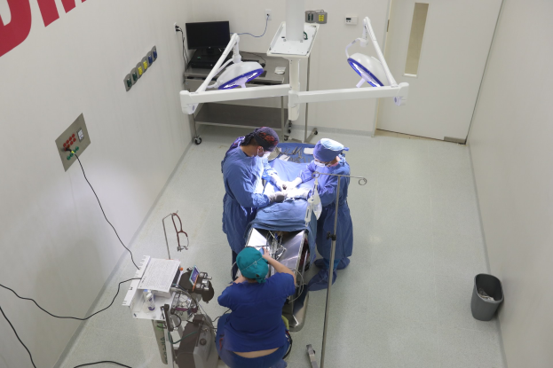 Eine Gruppe von Chirurgen führt einen Eingriff in einem Operationssaal durch, mit einem Patienten auf einer Trage umgeben von medizinischer Ausrüstung, einer textbedeckten Wand auf der linken Seite, einem Objekt auf dem Boden auf der rechten Seite und verschiedenen medizinischen Geräten im Hintergrund.