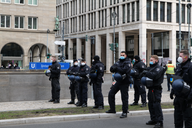 Gruppe von Polizisten in schwarzen Uniformen und Masken vor einem Berliner Gebäude mit Glasfenstern und Säulen, einige halten Helme, mit Laternenmästen, Verkehrsampeln, Schildern mit Text, einer Statue und anderen Menschen im Hintergrund.