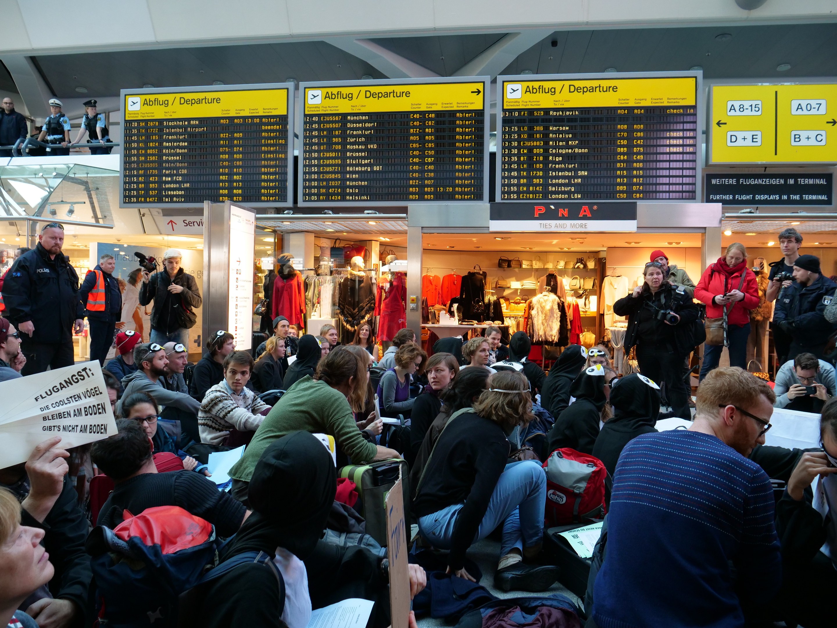 Eine große Gruppe von Menschen in einem Flughafen, einige sitzen mit Taschen und Papieren, andere stehen, mit Texttafeln, Schaufensterpuppen in Kleidern und Deckenbeleuchtung im Hintergrund, was auf eine Demonstration hindeutet.